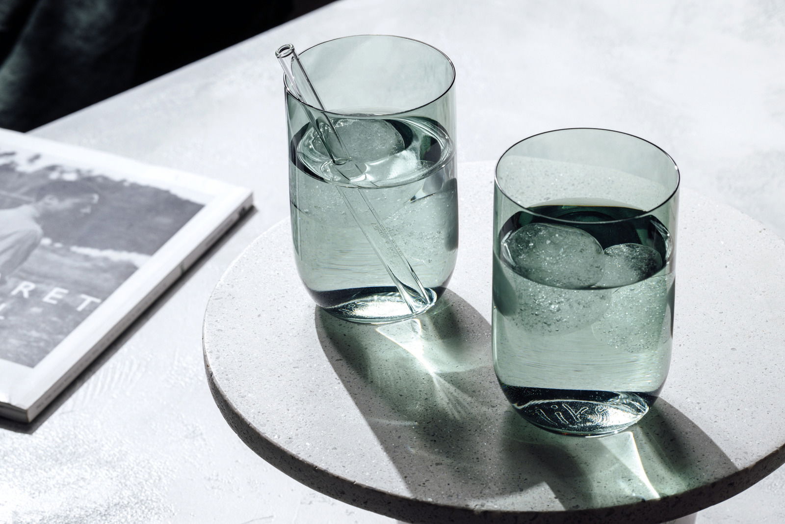 Zwei grüne Longdrinkbecher mit Eiswürfeln und einem Glasstrohhalm, auf einem runden, hellen Untersetzer von oben fotografiert.