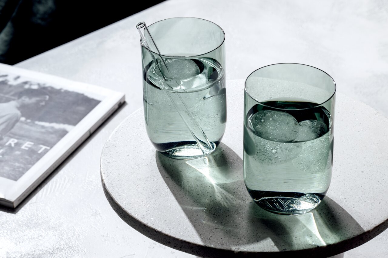 Zwei grüne Longdrinkbecher mit Eiswürfeln und einem Glasstrohhalm, auf einem runden, hellen Untersetzer von oben fotografiert.