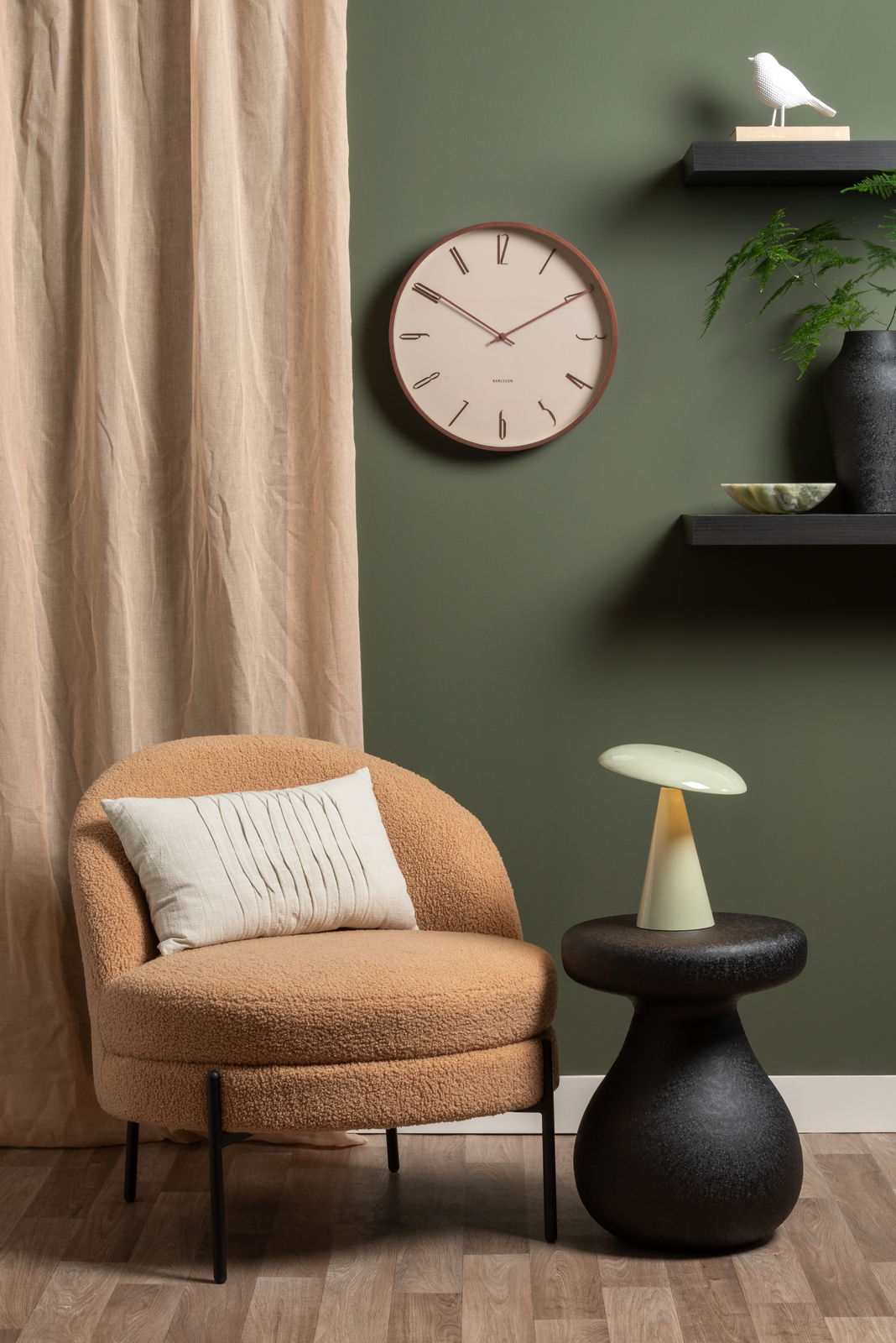 LED Akku Tischleuchte auf einem schwarzen Beistelltisch, neben einem beigen Sessel mit Kissen, vor einer grünen Wand mit Uhr und Regal. Perspektive: Frontalansicht.