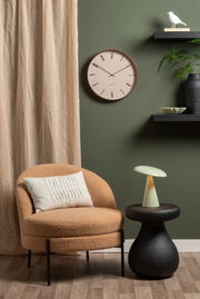 LED Akku Tischleuchte auf einem schwarzen Beistelltisch, neben einem beigen Sessel mit Kissen, vor einer grünen Wand mit Uhr und Regal. Perspektive: Frontalansicht.
