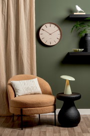 LED Akku Tischleuchte auf einem schwarzen Beistelltisch, neben einem beigen Sessel mit Kissen, vor einer grünen Wand mit Uhr und Regal. Perspektive: Frontalansicht.
