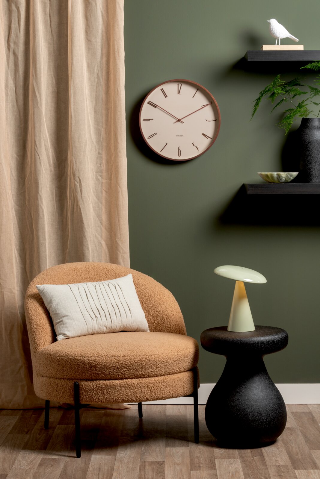 LED-tafellamp op een zwarte bijzettafel, naast een beige fauteuil met kussens, voor een groene muur met een klok en een plank. Perspectief: vooraanzicht.