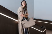 Frau in beigem Trenchcoat hält eine große, beige Tasche mit runden Griffen in einem modernen Innenraum. Perspektive: seitlich.