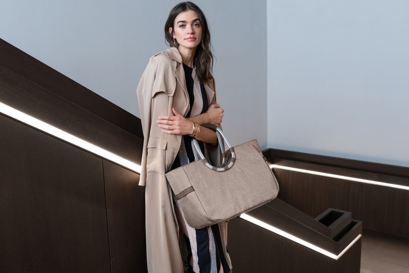 Frau in beigem Trenchcoat hält eine große, beige Tasche mit runden Griffen in einem modernen Innenraum. Perspektive: seitlich.