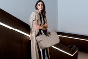 Frau in beigem Trenchcoat hält eine große, beige Tasche mit runden Griffen in einem modernen Innenraum. Perspektive: seitlich.