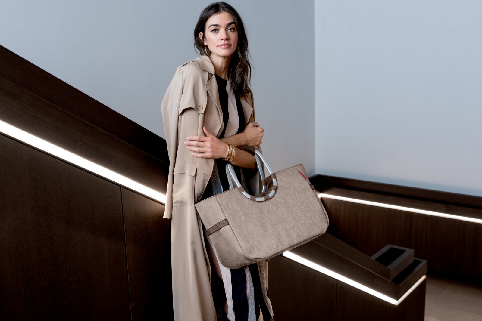 Een vrouw in een beige trenchcoat houdt een grote, beige tas met ronde handvatten vast in een modern interieur. Perspectief: zijaanzicht.