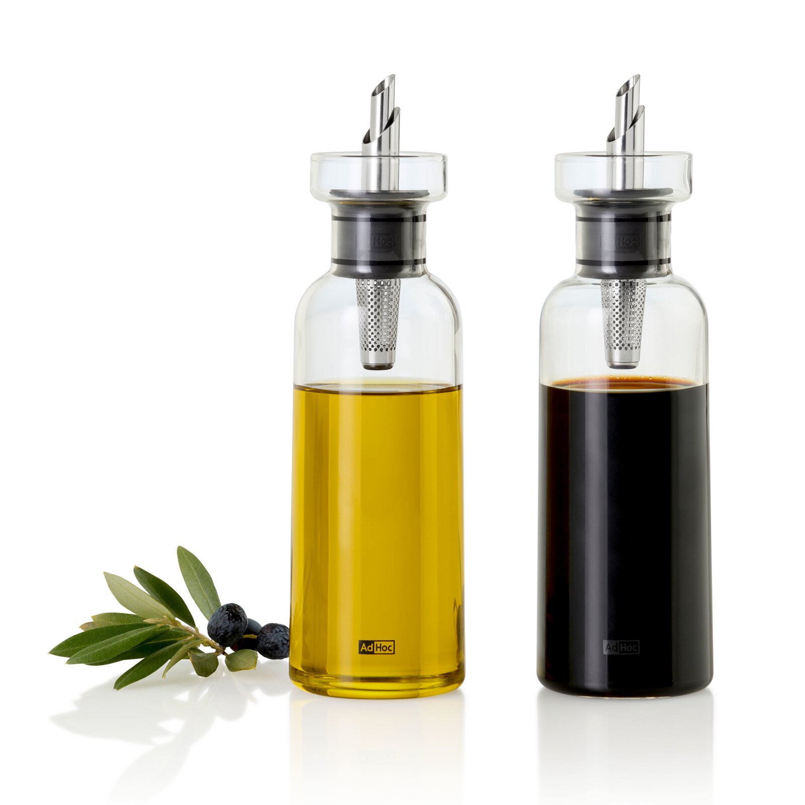 Zwei Glas-Öl- und Essigspender mit Edelstahl-Ausgießer und integriertem Filter, gefüllt mit goldenem Öl und dunklem Essig, frontale Produktansicht auf weißem Hintergrund; links Olivenzweig mit schwarzen Oliven.