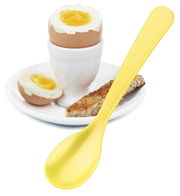 Gelber Eierlöffel in Nahaufnahme neben einem weichgekochten Ei im Eierbecher und einem Stück Toast auf einem weißen Teller.