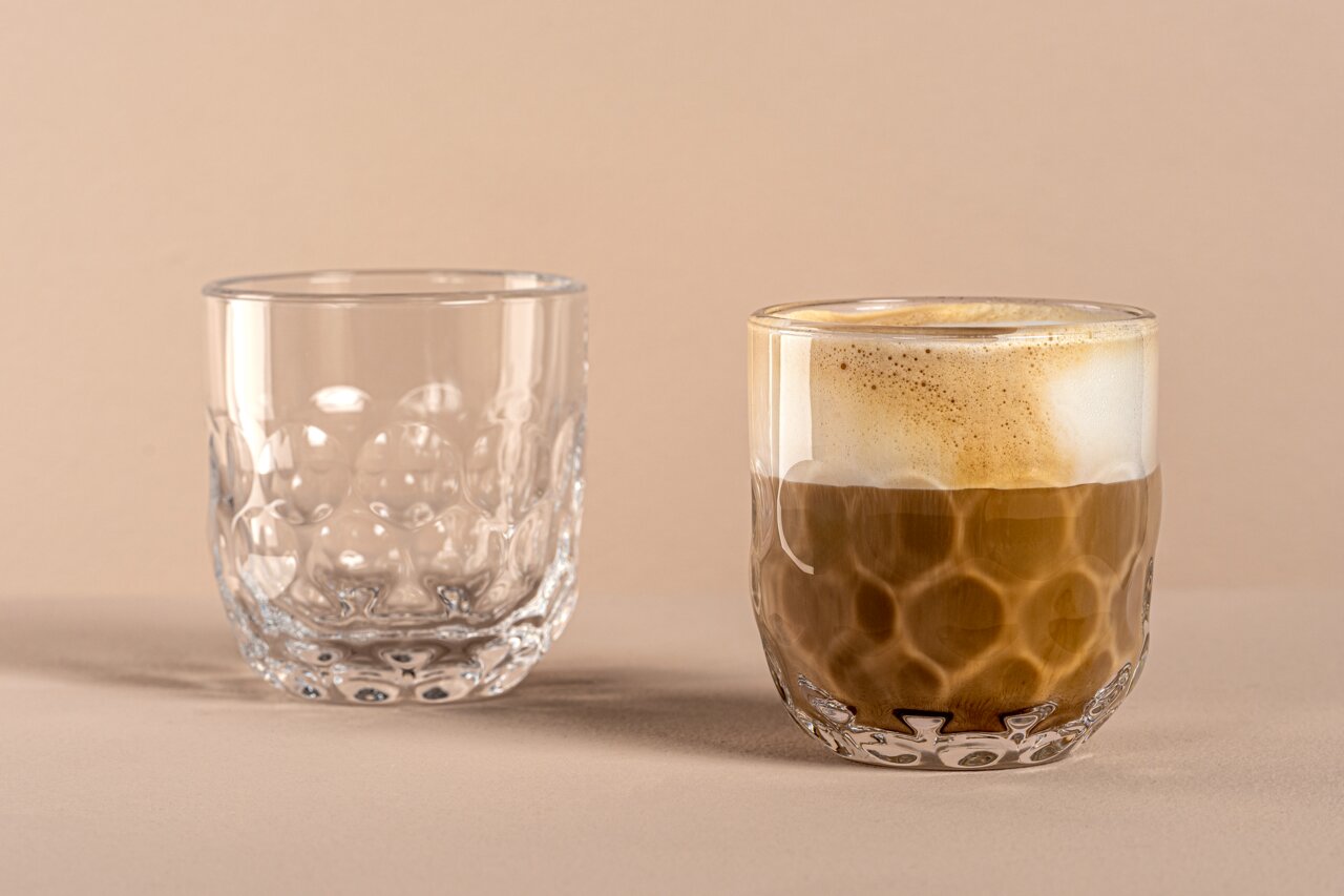 Zwei Gläser mit 220ml Volumen, eines leer und eines mit Kaffee gefüllt, auf einer neutralen Oberfläche. Die Gläser haben ein einzigartiges, tropfenförmiges Design. Perspektive von vorne.