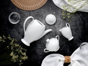 Draufsicht auf ein elegantes weißes Porzellan-Teeservice mit geriffelter Teekanne, Zuckerdose mit Deckel und Milchkännchen auf dunklem Steinuntergrund, dekoriert mit Stoffservietten, Blumen und einem Fächer.