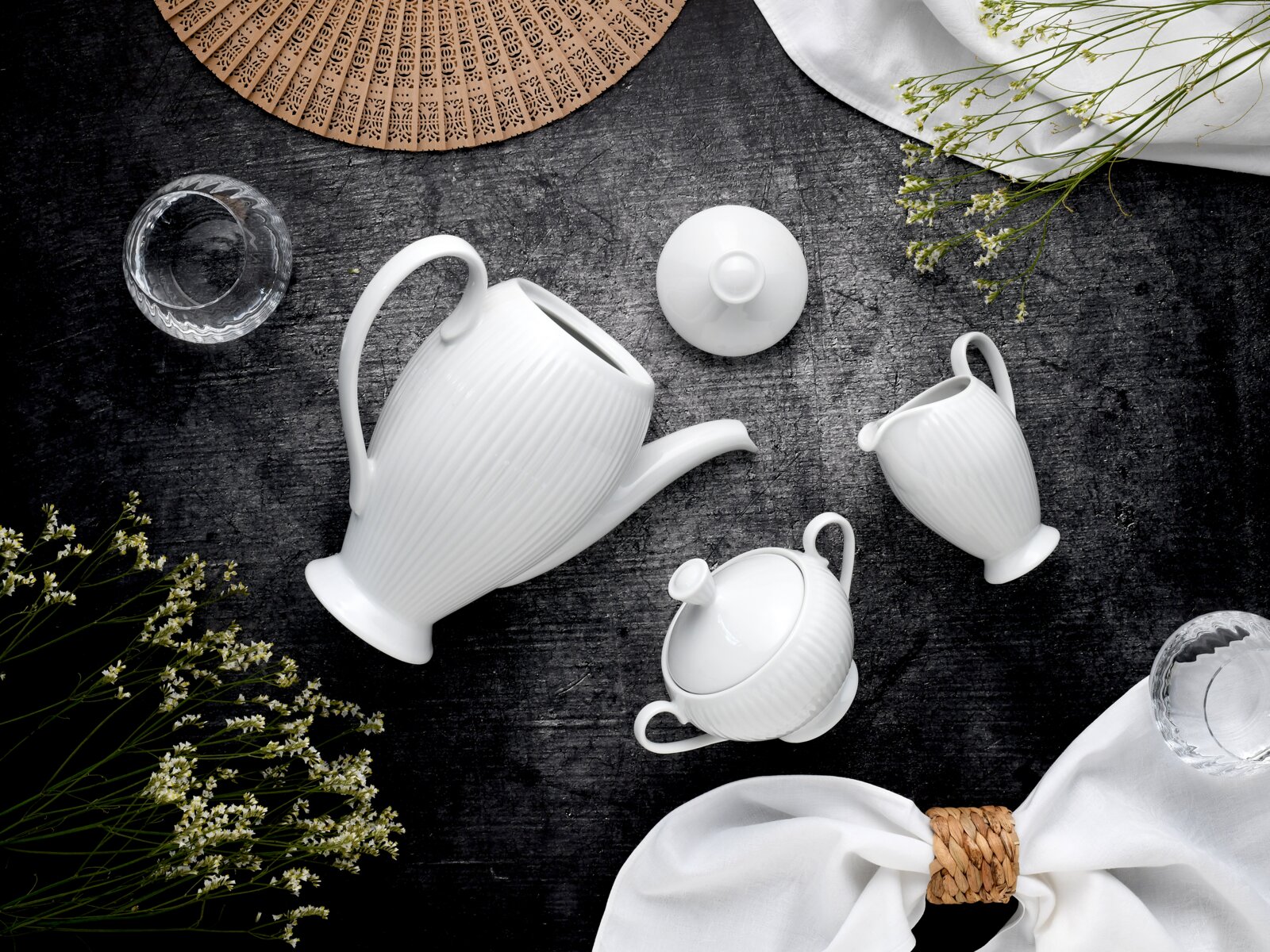 Draufsicht auf ein elegantes weißes Porzellan-Teeservice mit geriffelter Teekanne, Zuckerdose mit Deckel und Milchkännchen auf dunklem Steinuntergrund, dekoriert mit Stoffservietten, Blumen und einem Fächer.