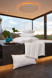 Ein Stapel von gesteppten Kopfkissen und Decken in einem modernen Raum mit großen Fenstern und Blick auf die Natur. Die Perspektive zeigt die Produkte von der Seite.