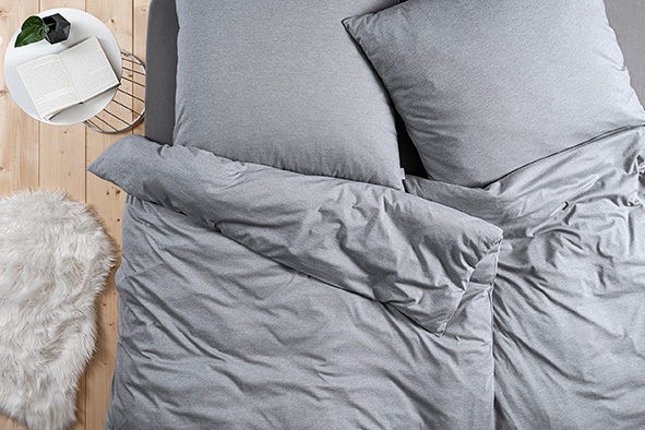 Graue Bettwäsche-Set bestehend aus einem Bettbezug 135x200 cm und einem Kissenbezug 80x80 cm, von oben fotografiert auf einem Holzboden mit einem runden Beistelltisch und einem weißen Teppich.