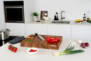 Modernes Küchen-Setting mit großem Holz-Schneidebrett auf der Arbeitsplatte, darauf Messer und rote Paprika; im Vordergrund Frühlingszwiebeln, Knoblauch, rote Zwiebeln, Tomaten und Schale mit Paprikastücken, Hintergrund mit Spüle und Backofen, Perspektive