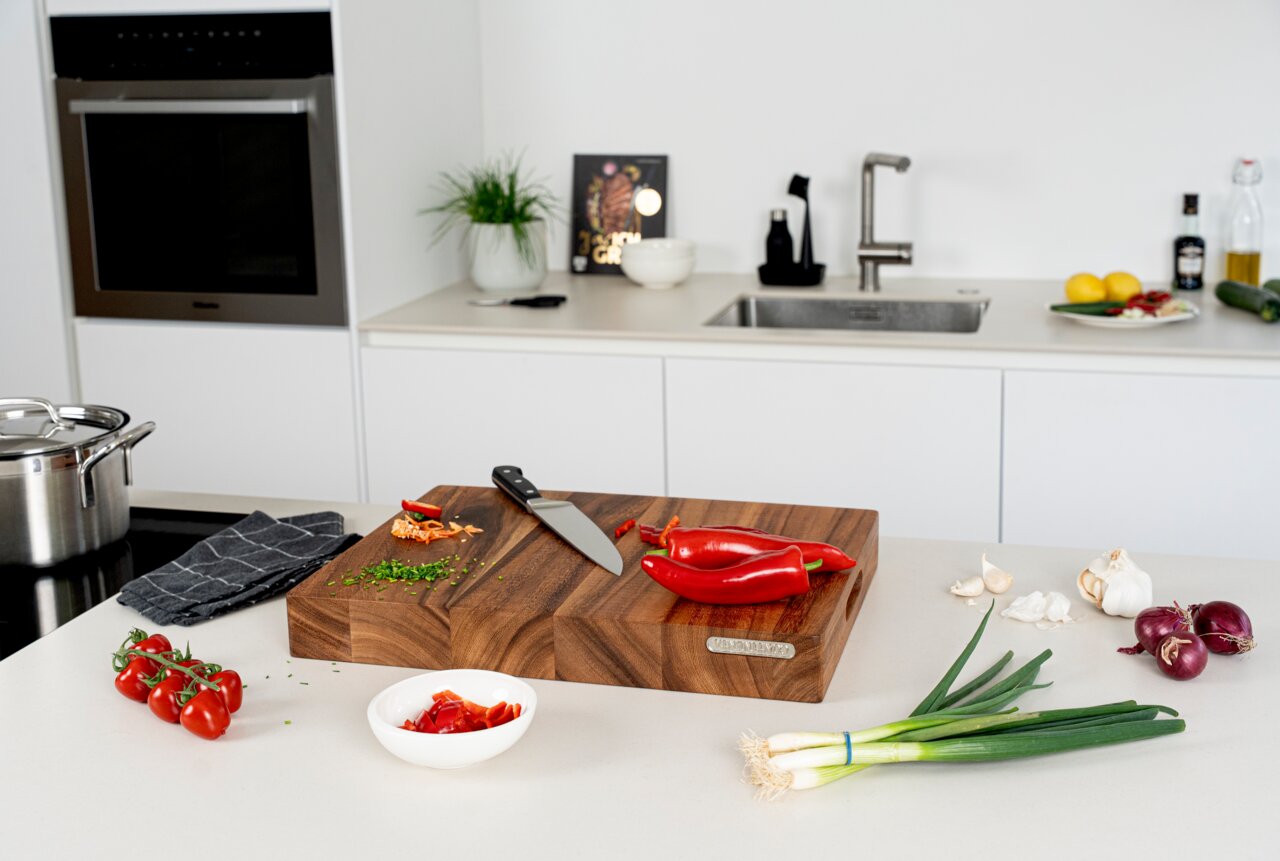 Modernes Küchen-Setting mit großem Holz-Schneidebrett auf der Arbeitsplatte, darauf Messer und rote Paprika im Vordergrund Frühlingszwiebeln, Knoblauch, rote Zwiebeln, Tomaten und Schale mit Paprikastücken, Hintergrund mit Spüle und Backofen, Perspektive