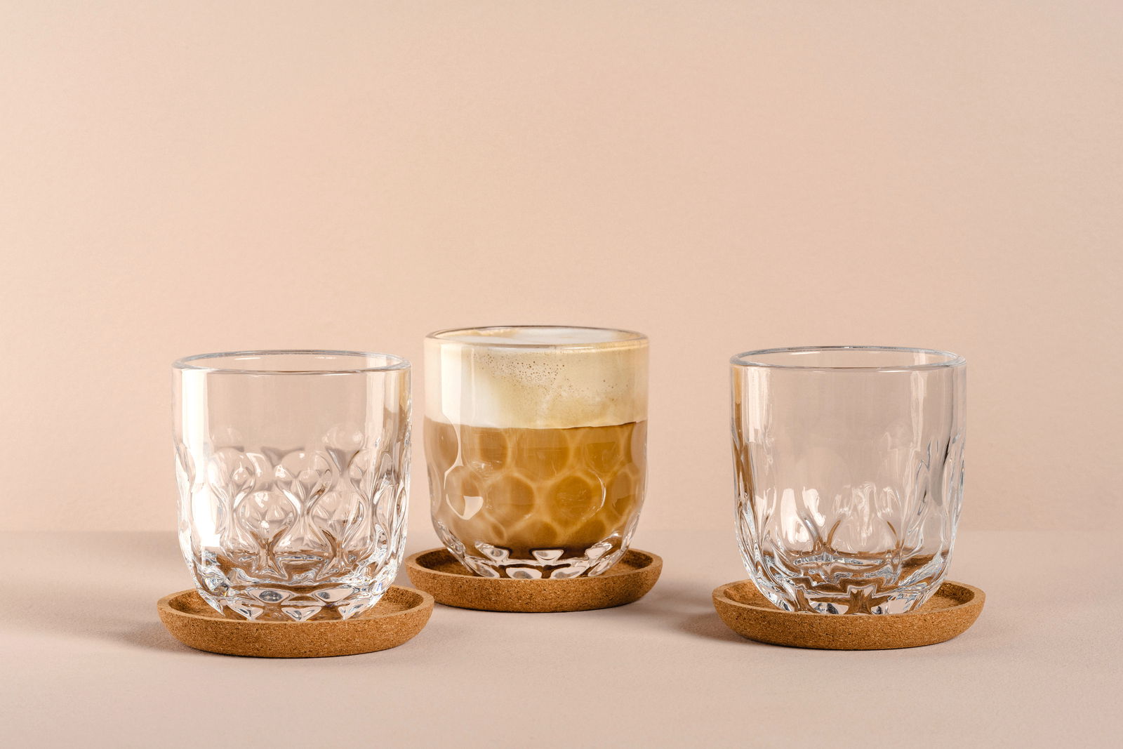 Drei elegante Gläser auf Korkuntersetzern, eines mit Kaffee gefüllt, aus frontaler Perspektive.