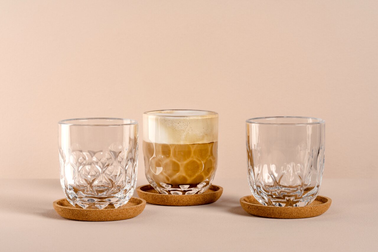 Drei elegante Gläser auf Korkuntersetzern, eines mit Kaffee gefüllt, aus frontaler Perspektive.