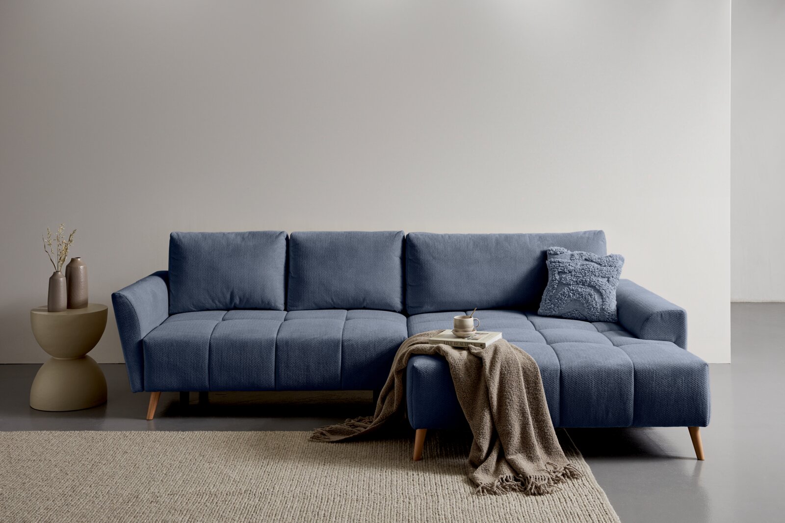 Z2 Ecksofa ZELDA Modernes blaues Ecksofa mit Chaise longue rechts, gestepptem Stoffbezug und Holzfüßen, frontal aus leicht schräger Perspektive fotografiert; dekoriert mit Kissen, Decke und Tablett im minimalistischen Wohnraum.