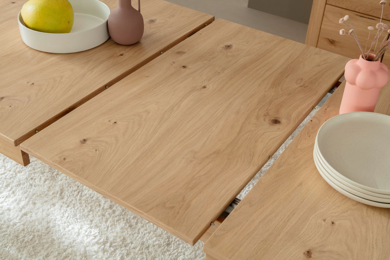 Nahaufnahme eines Esstisches aus Holz mit natürlicher Maserung, dekoriert mit einer Schale, einer Vase und Tellern, aus der Vogelperspektive.