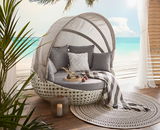 Gartenmuschel mit grauen Kissen und Decke, auf einer Terrasse mit Meerblick, aus seitlicher Perspektive fotografiert.