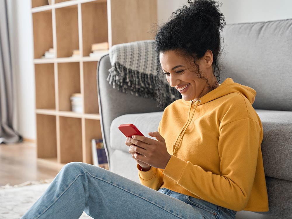 Een vrouw zit voor een grijze bank en kijkt naar haar mobiele telefoon.