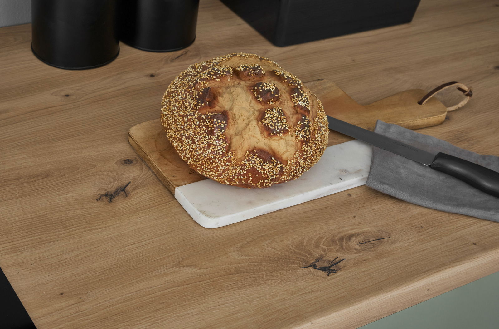 Nahaufnahme eines rustikalen Brotes mit Körnern auf einem Schneidebrett in einer modernen Küche, seitliche Perspektive.