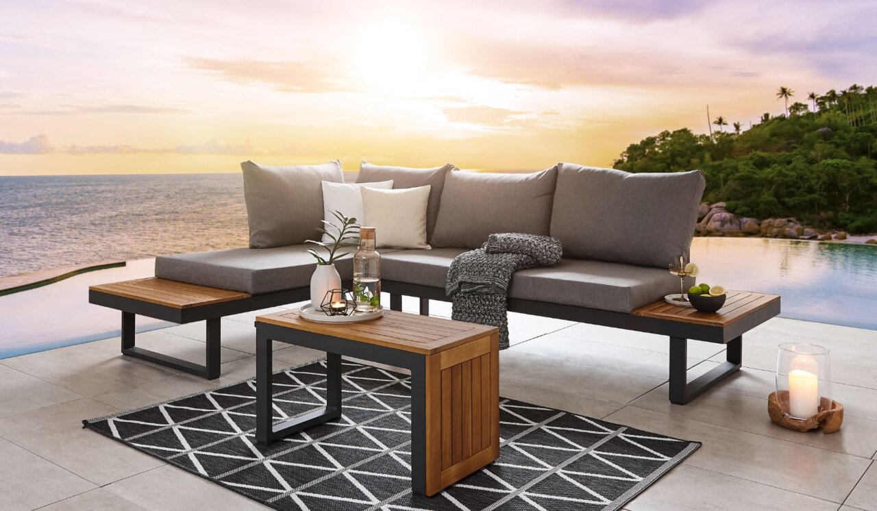 Moderne Gartenlounge mit grauen Kissen und Holzelementen, im Vordergrund ein Tisch mit Dekoration, im Hintergrund ein Pool und Sonnenuntergang, seitliche Perspektive.