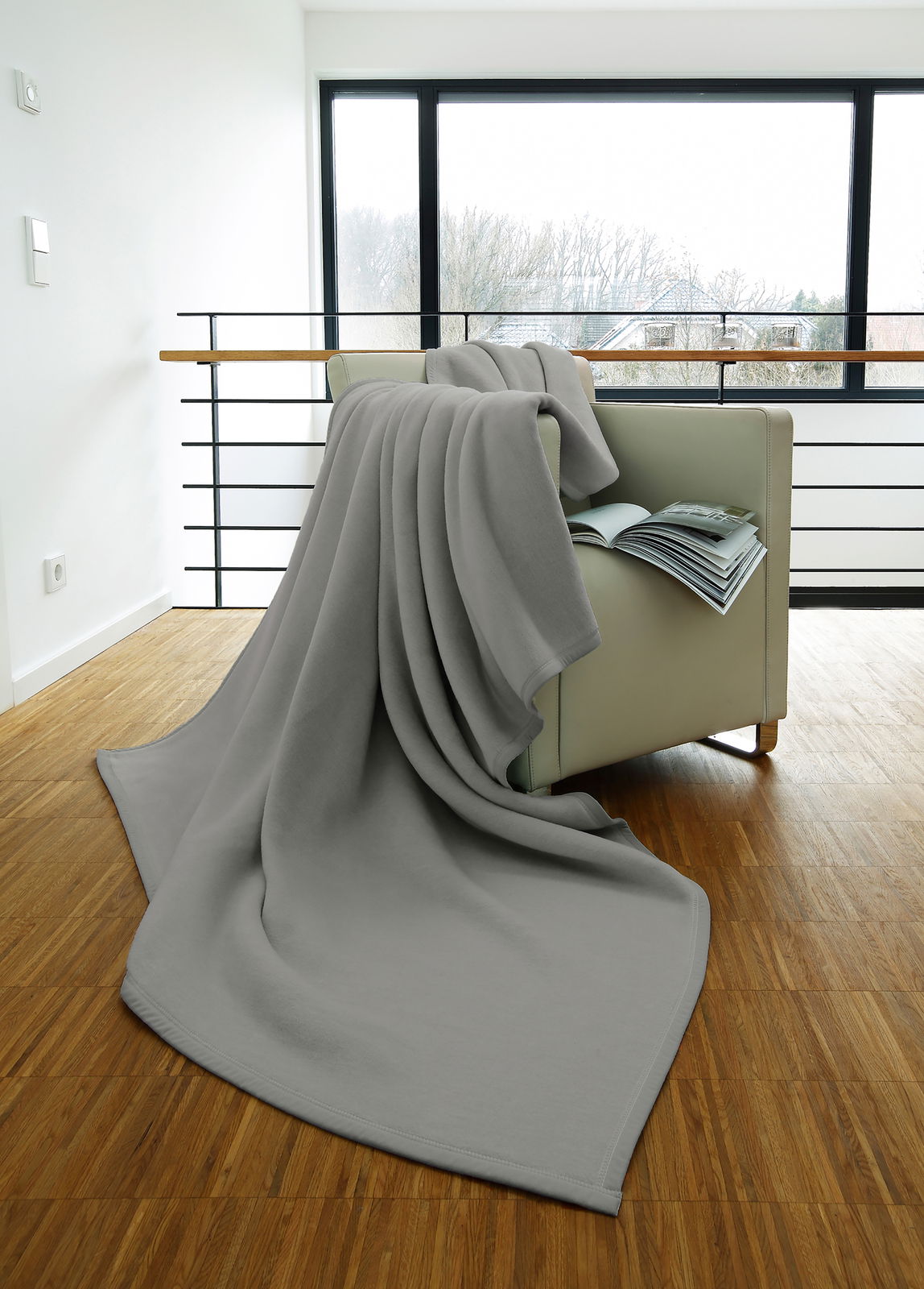 Graue Wohndecke 150 x 200 cm auf einem Sessel drapiert, mit Blick auf ein großes Fenster im Hintergrund.