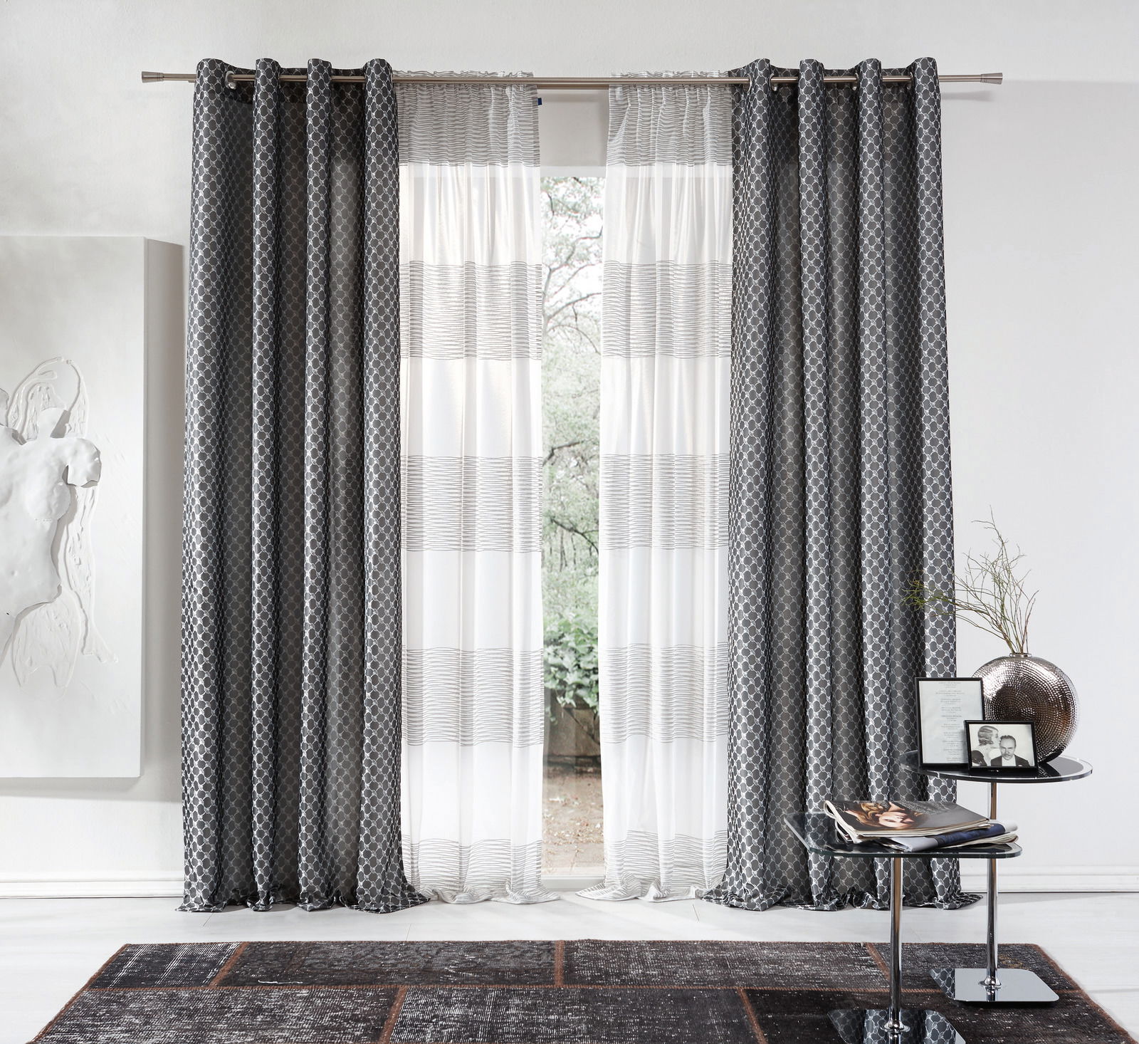 JOOP! Ösenvorhang grau Allovers Grauer Ösenvorhang von Stöckel & Grimmler, Modell JOOP, in einem modernen Wohnzimmer. Der Vorhang hat ein elegantes Muster und hängt an einer Metallstange. Die Perspektive zeigt den Vorhang frontal mit einem Blick nach draußen durch das Fenster.