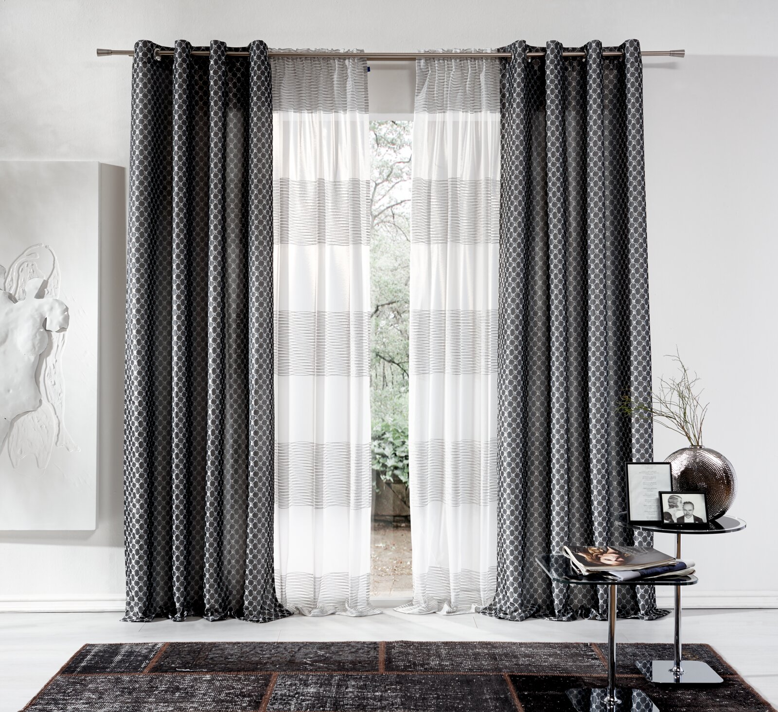 Grauer Ösenvorhang von Stöckel & Grimmler, Modell JOOP, in einem modernen Wohnzimmer. Der Vorhang hat ein elegantes Muster und hängt an einer Metallstange. Die Perspektive zeigt den Vorhang frontal mit einem Blick nach draußen durch das Fenster.
