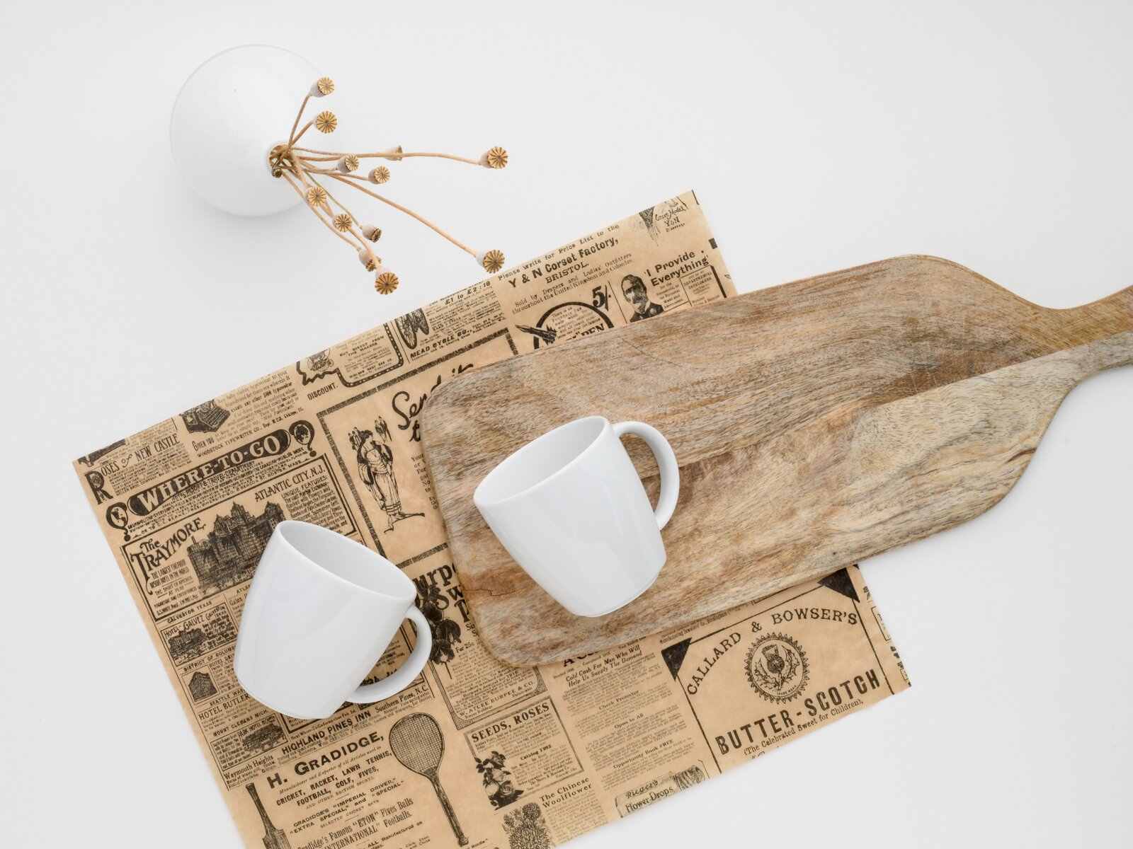 Rustikales Holz-Servierbrett mit Griff auf Vintage-Zeitungspapier, daneben zwei weiße Tassen; Draufsicht (Flatlay) auf hellem Hintergrund.