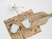 Rustikales Holz-Servierbrett mit Griff auf Vintage-Zeitungspapier, daneben zwei weiße Tassen; Draufsicht (Flatlay) auf hellem Hintergrund.