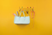 Bunte Garderobe mit Haken und Kugeln, an der eine Tasche, Geschenkpapierrollen, eine Schere und ein Seil hängen, vor gelbem Hintergrund, frontal aufgenommen.