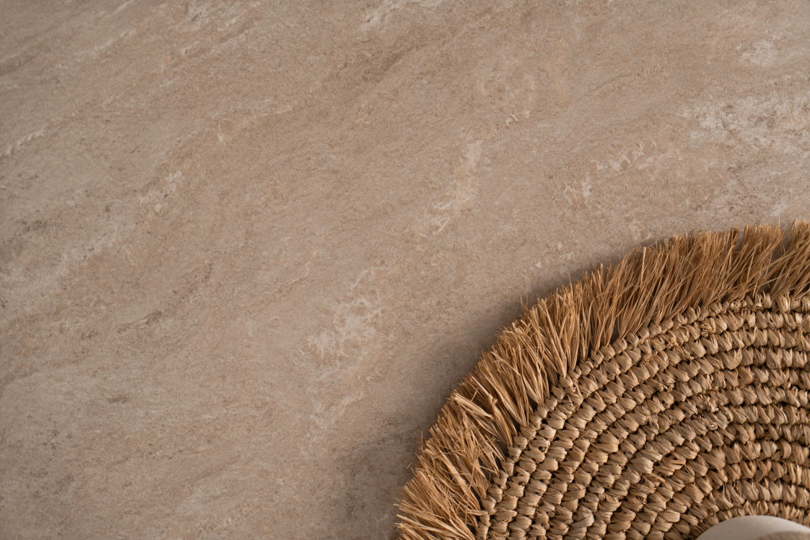 Nahaufnahme eines Esstisches mit natürlicher Steinoptik und einem runden, geflochtenen Tischset aus Stroh, von oben betrachtet.