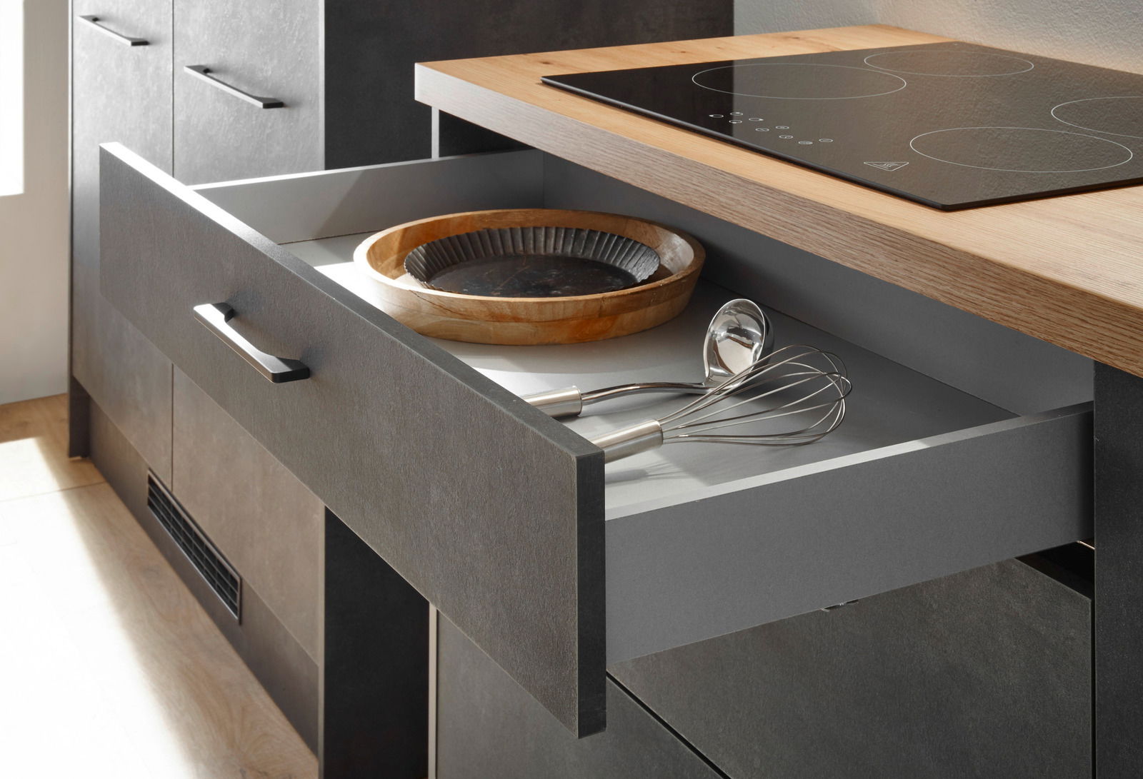 Geöffnete Küchenschublade mit Schneebesen und Schöpfkelle, darüber eine Arbeitsplatte mit Induktionskochfeld, seitliche Perspektive.