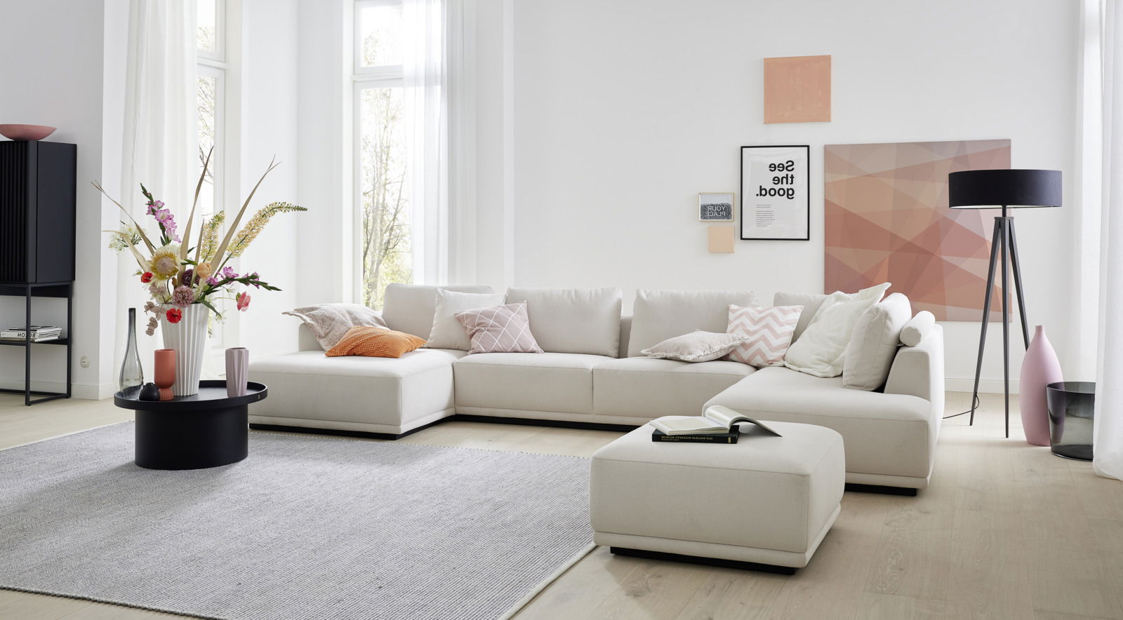 Moderne Polsterrundecke in einem hellen Wohnzimmer, mit beigen Kissen und einem passenden Hocker, aus der Perspektive von vorne fotografiert.