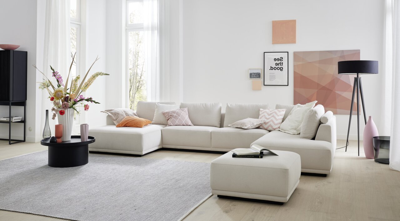 Een moderne gestoffeerde hoekbank in een lichte woonkamer, met beige kussens en een bijpassende poef, gefotografeerd vanuit het vooraanzicht.