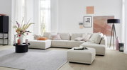 Moderne Polsterrundecke in einem hellen Wohnzimmer, mit beigen Kissen und einem passenden Hocker, aus der Perspektive von vorne fotografiert.