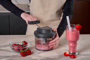Persoon gebruikt een handmatige hakmolen met trekkoord om aardbeien tot een roze smoothie te mengen op een marmeren aanrecht, met een glas aardbeiensmoothie en verse bessen ernaast, gefotografeerd in vooraanzicht op werkbladhoogte.