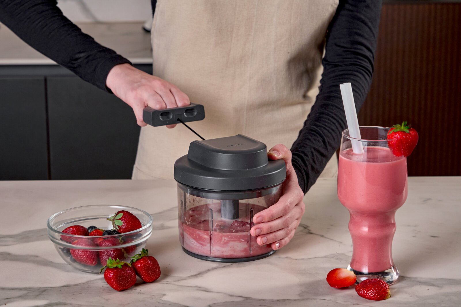 Persoon gebruikt een handmatige hakmolen met trekkoord om aardbeien tot een roze smoothie te mengen op een marmeren aanrecht, met een glas aardbeiensmoothie en verse bessen ernaast, gefotografeerd in vooraanzicht op werkbladhoogte.
