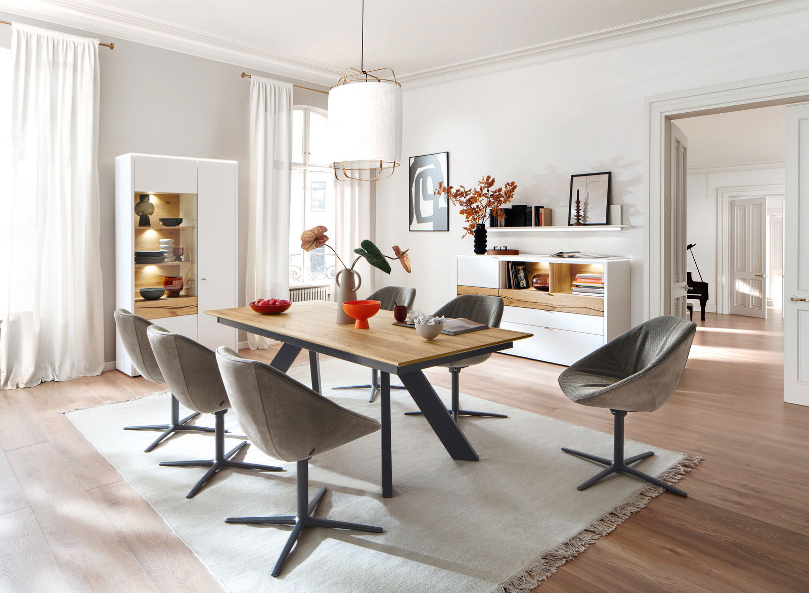 Esszimmer mit modernem Esstisch aus Holz und Metall, umgeben von grauen Stühlen auf einem hellen Teppich. Im Hintergrund eine beleuchtete Vitrine und ein Sideboard, seitliche Perspektive.