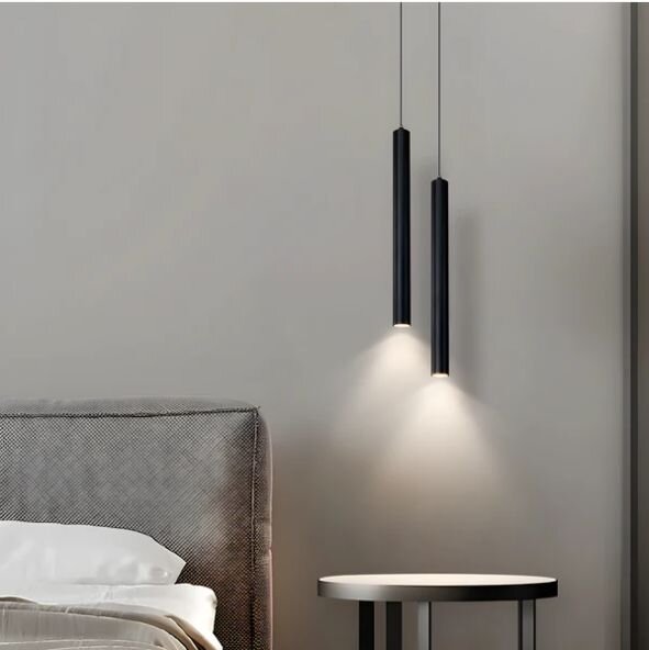 Zwei schlanke schwarze zylindrische Pendelleuchten hängen neben dem Bett und werfen warmes Licht auf eine graue Wand, seitliche Perspektive.