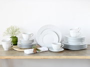 Weißes Porzellan-Geschirrset mit Tellern und Tassen auf Holztisch arrangiert, dekoriert mit grünem Vase-Blumenschmuck und Serviette; frontale Ansicht auf Augenhöhe.