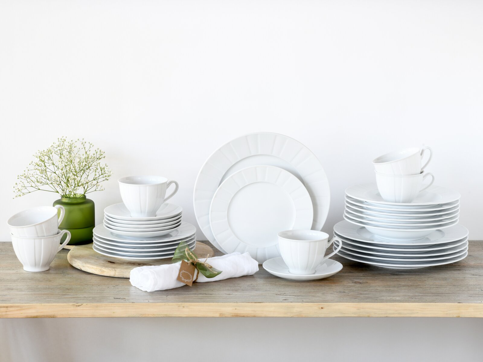 Weißes Porzellan-Geschirrset mit Tellern und Tassen auf Holztisch arrangiert, dekoriert mit grünem Vase-Blumenschmuck und Serviette; frontale Ansicht auf Augenhöhe.