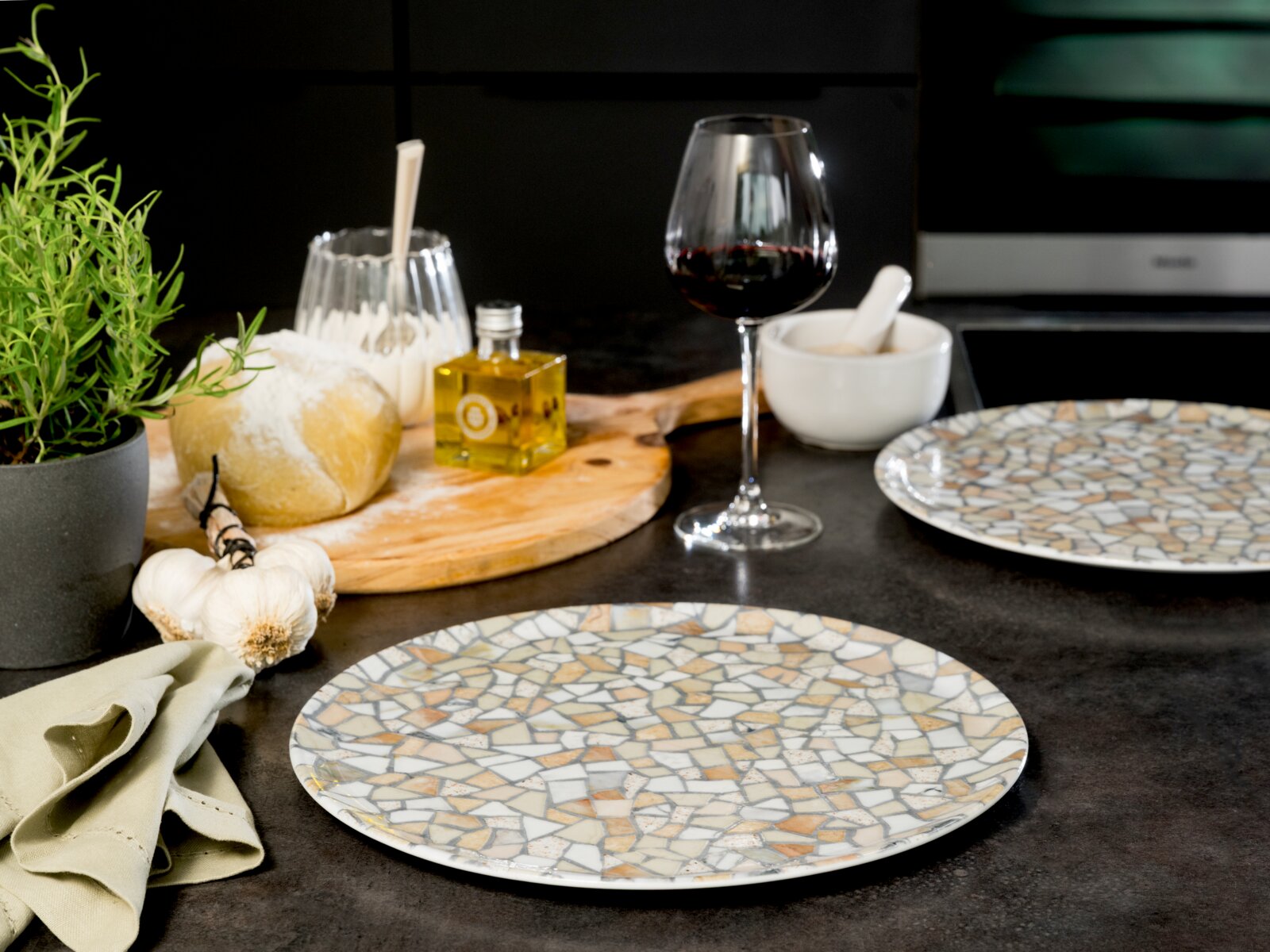 Zwei flache Speiseteller mit Mosaikmuster in Grau-, Beige- und Weißtönen auf dunkler Küchenarbeitsplatte, Vordergrund- und leicht seitliche Perspektive; im Hintergrund Brot, Olivenöl, Rotweinglas und Küchenutensilien.
