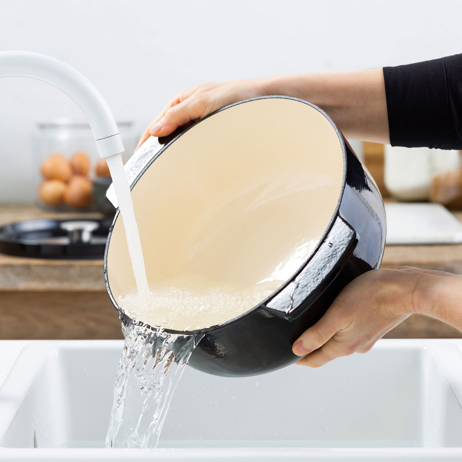Gusseiserner ovaler Bräter mit cremefarbener Innenbeschichtung, der in einem Waschbecken unter fließendem Wasser gereinigt wird. Perspektive von der Seite.