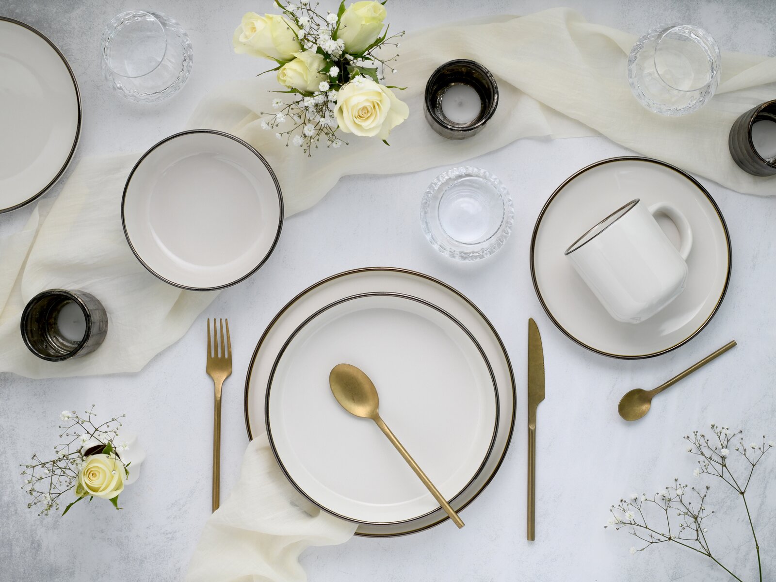 Draufsicht auf ein elegant gedecktes Tischarrangement mit weißen Tellern und Tasse mit dunklem Rand, goldfarbenem Besteck, Glas-Schälchen, Kerzenhaltern und weißen Rosen auf hellem Tuch.