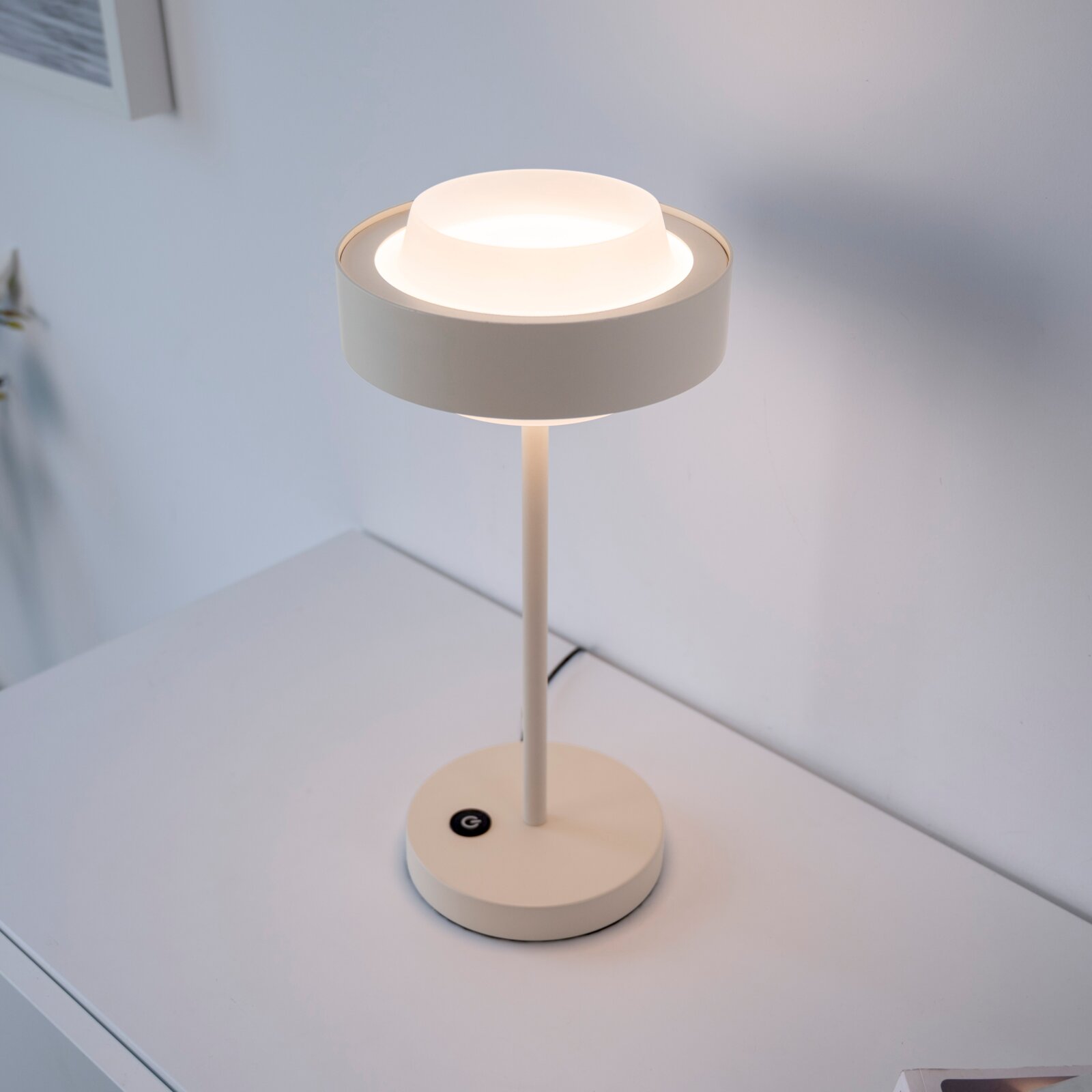 Moderne weiße LED-Tischlampe mit rundem Sockel und zylindrischem Schirm auf einem weißen Tisch, leuchtend, fotografiert aus leicht erhöhter schräger Vorderansicht