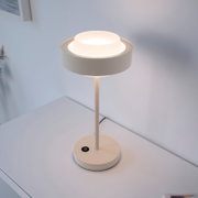 Moderne weiße LED-Tischlampe mit rundem Sockel und zylindrischem Schirm auf einem weißen Tisch, leuchtend, fotografiert aus leicht erhöhter schräger Vorderansicht
