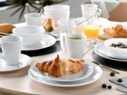 Helles Frühstücks-Table-Setting mit weißem Porzellan-Geschirr, Kaffeetassen, Schalen und Tellern, Croissants, Orangensaft, Wasser, Obst und Blaubeeren auf Holztisch, aufgenommen aus leicht erhöhter schräger Perspektive